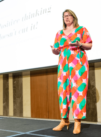 The photo shows a woman in an orange jumpsuit talking to an audience. 